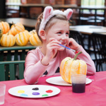 A child wearing a white animal-ear headband sits at a craft table painting a small striped pumpkin. The table is covered with a pink tablecloth and holds paint palettes, brushes and cups of water. Numerous decorative pumpkins are arranged on tables in the background, suggesting a seasonal arts and crafts activity in an indoor community or event space.