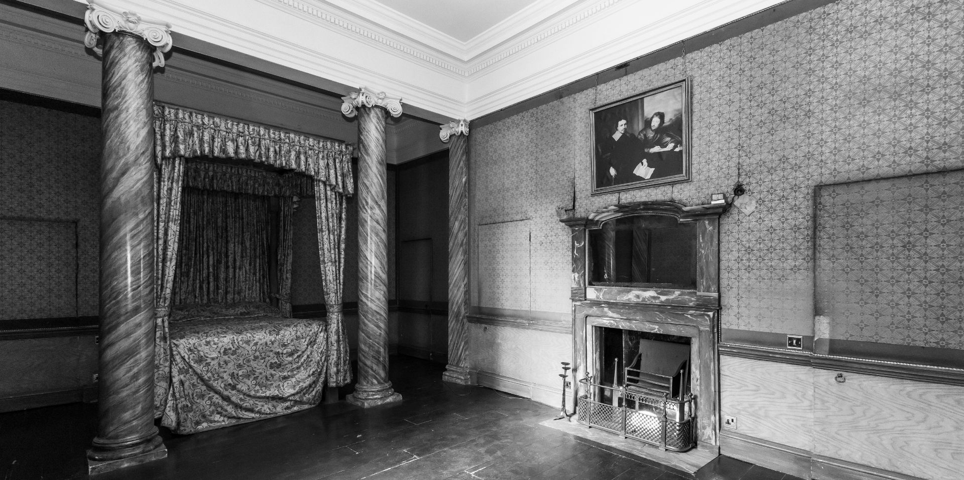Black‑and‑white photo of an old-fashioned bedroom with a large four‑poster bed framed by two ornate columns. Opposite the bed is a decorative fireplace with a metal fireguard, and a framed portrait hanging above it. The room has patterned wallpaper, high ceilings with detailed cornicing, and an overall historic, slightly worn appearance.