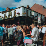 A busy street scene outside the Frog & Parrot pub on a sunny day, with large crowds gathered around the historic building decorated with hanging flower baskets.