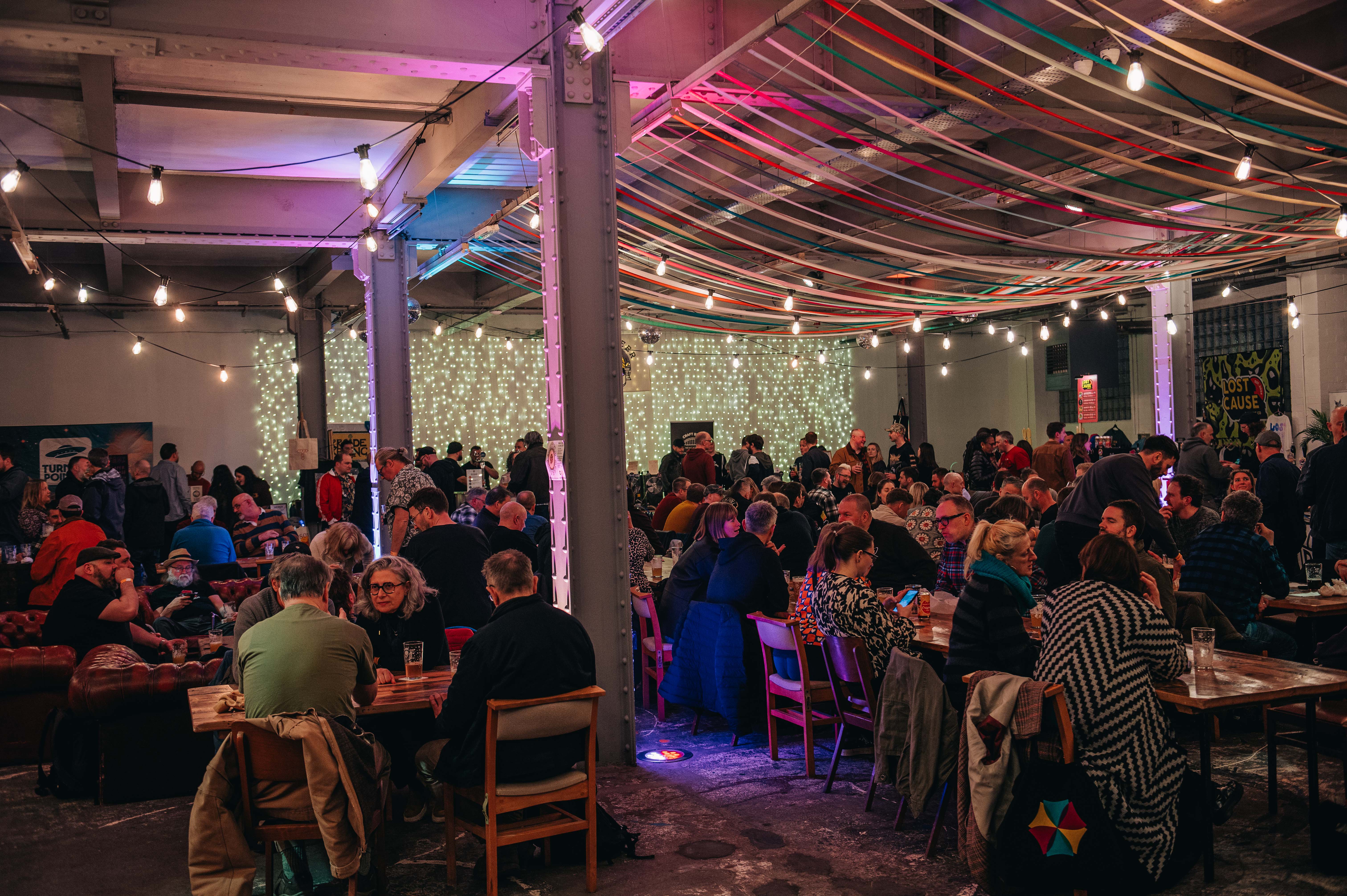 Wide view of a lively indoor beer festival with rows of tables filled with people, illuminated by strings of lights and ambient coloured lighting.