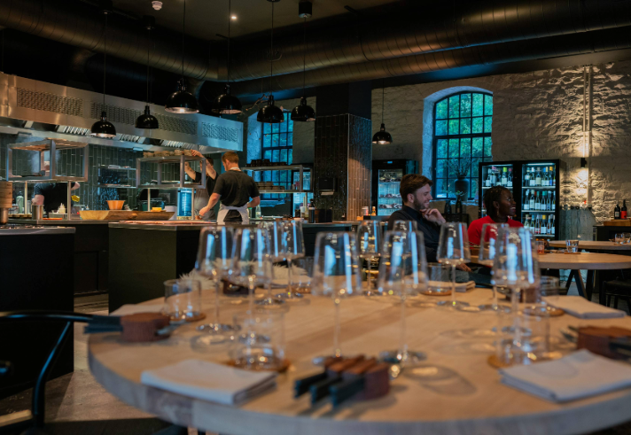 A modern, stylish restaurant interior featuring an open kitchen with a chef at work. In the foreground, a round wooden table is set with multiple empty wine glasses, folded napkins, and cutlery. The space has exposed stone walls, large arched windows, and shelves stocked with bottles. Black pendant lights hang from the ceiling, and the atmosphere is warm and contemporary.