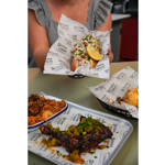 A tabletop spread of seafood and barbecue dishes at Butcher & Catch. In the foreground, a glazed grilled chicken leg topped with chopped pickles and herbs sits on a tray lined with branded paper. A baked dish and a loaded roll are nearby. In the background, a person presents a lobster roll packed with seafood salad, garnished with herbs and a lemon wedge. The restaurant interior is softly blurred behind the food.