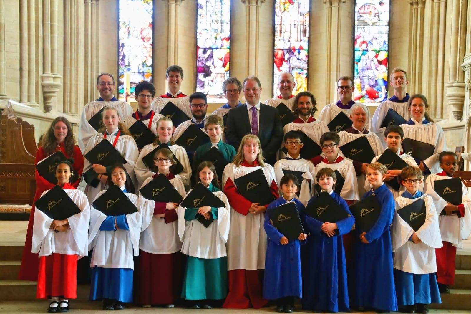 A large choir group dressed in traditional robes stands inside a grand cathedral. The robes are in various colors, including white, red, blue, and green, and many members hold black folders with gold detailing. Behind the group are tall stone columns and vibrant stained-glass windows that fill the background with colorful light. The setting appears formal and historic, emphasising the architectural details and the ceremonial attire of the choir.