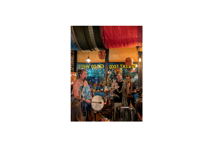 Live music performance inside a cosy café decorated with colourful hanging fabrics, flowers and warm lighting. Three performers stand with microphones and instruments, including a hand drum and acoustic guitar, in front of a window painted with the words “Great Food” and “Good Vibes.” Audience members are visible in the foreground, while decorative branches and lamps create an intimate, bohemian atmosphere.