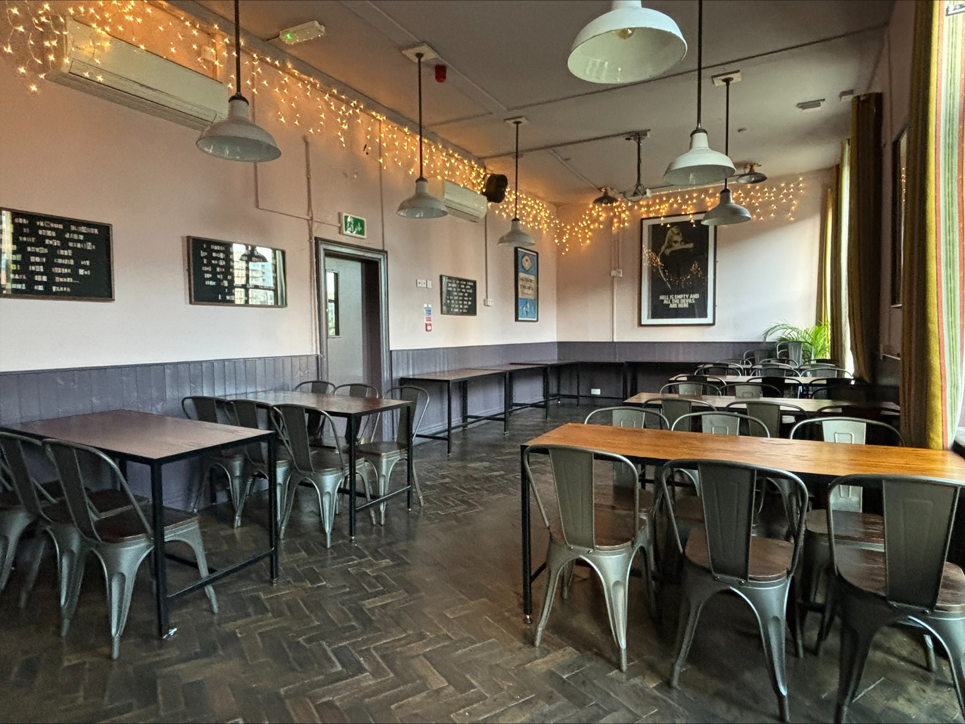 A cosy room with wooden floors, metal chairs and simple tables arranged in rows. Warm string lights hang across the ceiling, with framed boards and posters on the walls. Large curtained windows let in natural light, creating a relaxed event or meeting space.