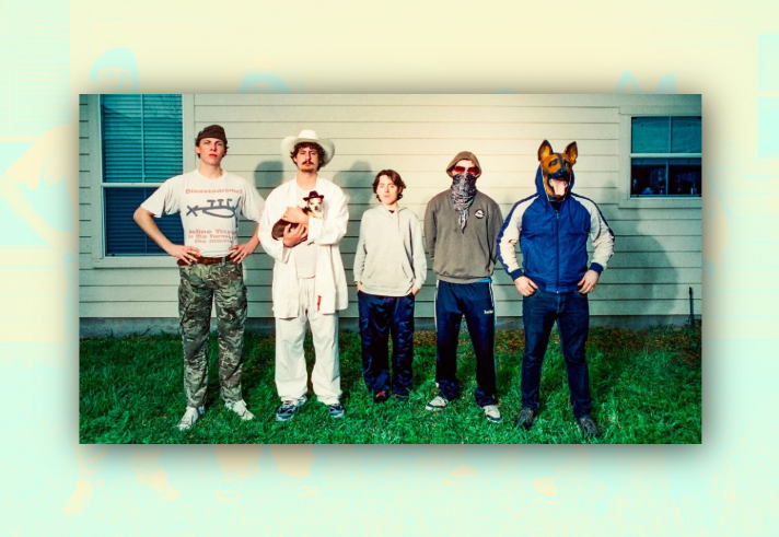 Five people stand in a line on grass in front of a light-coloured building wall, each wearing distinctive outfits. One wears camouflage trousers and a graphic T-shirt, another wears a white cowboy hat and holds a small dog, a third wears a plain hoodie, a fourth has a hood, sunglasses and a face covering, and the fifth wears a blue tracksuit top with a dog mask. The image has a quirky, staged band-promo style with strong sunlight and long shadows.