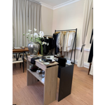 Interior of a minimalist boutique or showroom with cream walls, tall curtains and wooden flooring. In the foreground, a display table holds a large silver vase filled with white flowers alongside trays, dishes and accessories. Behind it, two clothing rails display neatly arranged garments in neutral shades including white, beige, grey and black. A wooden table and chairs sit to the left, creating a calm, curated retail setting.