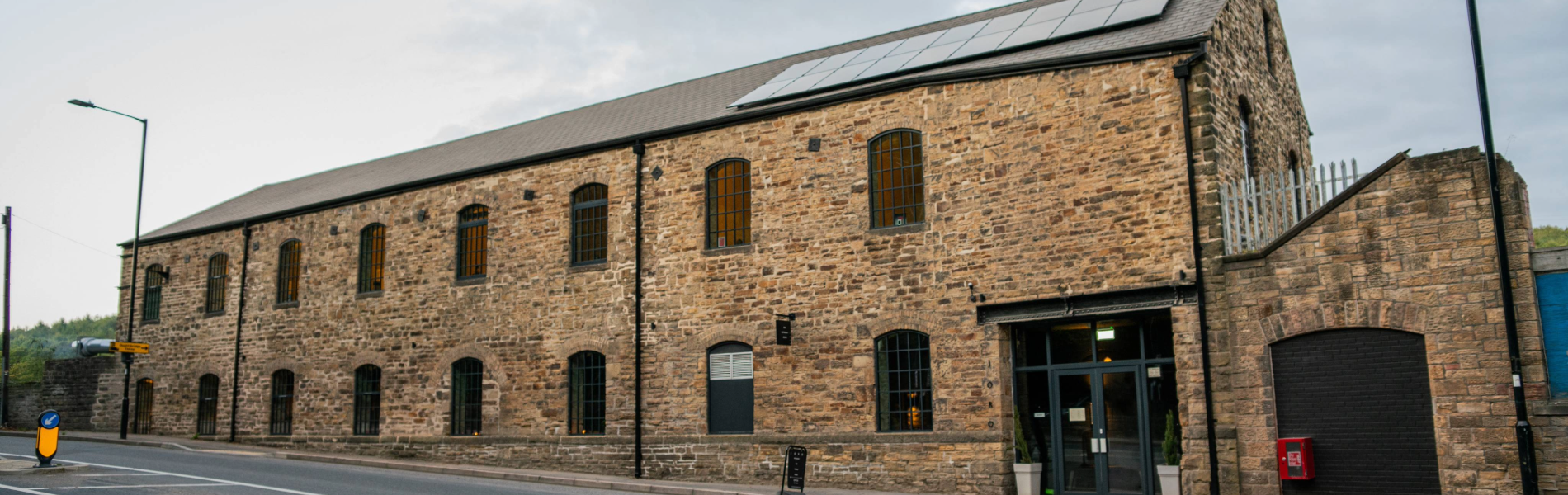 Exterior view of a large stone building with tall arched windows and a pitched roof fitted with solar panels. The structure has an industrial yet modern look, with a glass entrance door flanked by potted plants. It sits alongside a paved road with white markings, and the sky above is overcast. The building appears to be a converted mill or warehouse with contemporary features.