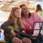 A young girl sits holding a guitar-style video game controller while smiling at a screen out of view. Beside her, an adult woman in a pink jumper smiles and gestures encouragingly. A music stand with sheet music is in the foreground. The scene takes place in a brightly lit indoor venue with blurred arcade machines and visitors in the background, suggesting a family-friendly gaming or interactive exhibition space.