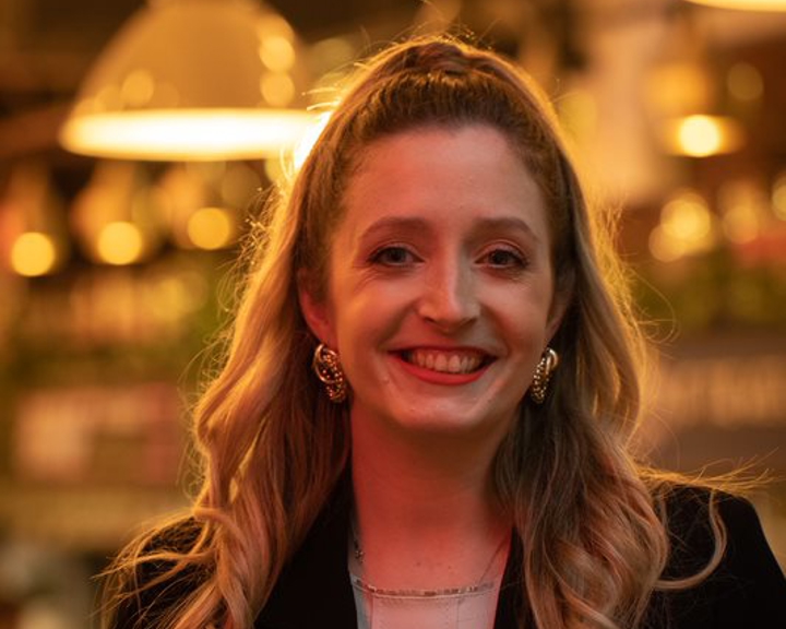 Person with long wavy hair wearing a light-colored top and a dark jacket, standing indoors under warm ambient lighting with hanging lamps and blurred background decor.