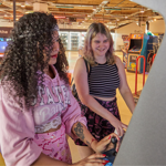 Two young people play an arcade machine together in a bright gaming museum or arcade space. One person in a pink hoodie focuses on the controls while the other smiles as they watch the screen. Classic arcade cabinets and interactive exhibits are visible in the background, creating a lively atmosphere. The image captures the enjoyment and social experience of playing video games together.