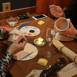 People painting ceramic mugs at a table with paint palettes, brushes, candles, and drinks during a pottery painting session.