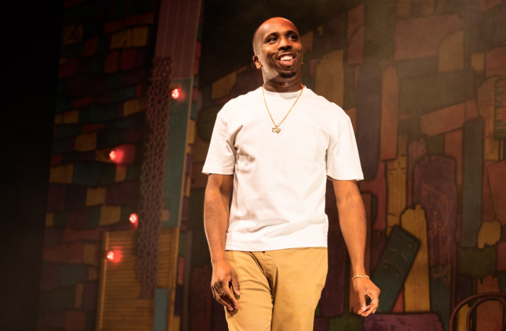 Person wearing a plain white short-sleeved T-shirt, beige trousers, and gold jewellery including a necklace and bracelet, standing on a stage. The background features a colourful, abstract mural with geometric shapes in warm tones and several small red lights. The scene is warmly lit, creating a theatrical atmosphere.