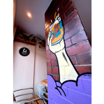 Interior view of Abbeydale Beerworks Taproom. A painted mural covers a red brick pillar, showing a large hand holding a foaming beer glass filled with colourful illustrations of books, buildings and other objects. The lower section of the pillar is painted purple. In the background are wooden shelves with potted plants, metal-framed chairs, and an Abbeydale Brewery wall sign. Warm ceiling lighting illuminates the modern taproom space.