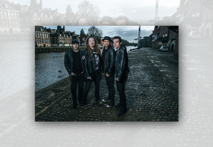 Four people dressed in dark clothing and leather jackets stand on a cobbled riverside quay beside a waterway lined with historic brick buildings. One person wears a flat cap, another has long dreadlocks, and a third wears a bowler-style hat. The overcast sky and wet cobbles create a moody urban atmosphere, with a bridge visible in the distance across the river.