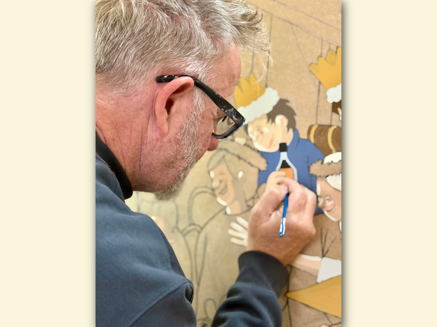 A person is painting a detailed illustration on a textured surface. The artwork features cartoon-style characters wearing festive crowns and fur-trimmed outfits, gathered around a table. One character is holding a bottle, and the scene suggests a celebratory or holiday setting. The painter’s hand is visible holding a fine brush, adding colour to the design with precision.