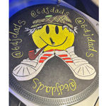 Close-up of a customised DJ turntable slipmat featuring a large yellow smiley face wearing a patterned bucket hat, smoking a pipe, and resting on a pair of white trainers with red stripes. Handwritten yellow text curves around the design, reading “@6djdads” and “@6djdas”. The circular artwork sits on a black turntable platter with metallic edging, illuminated by blue-purple lighting.