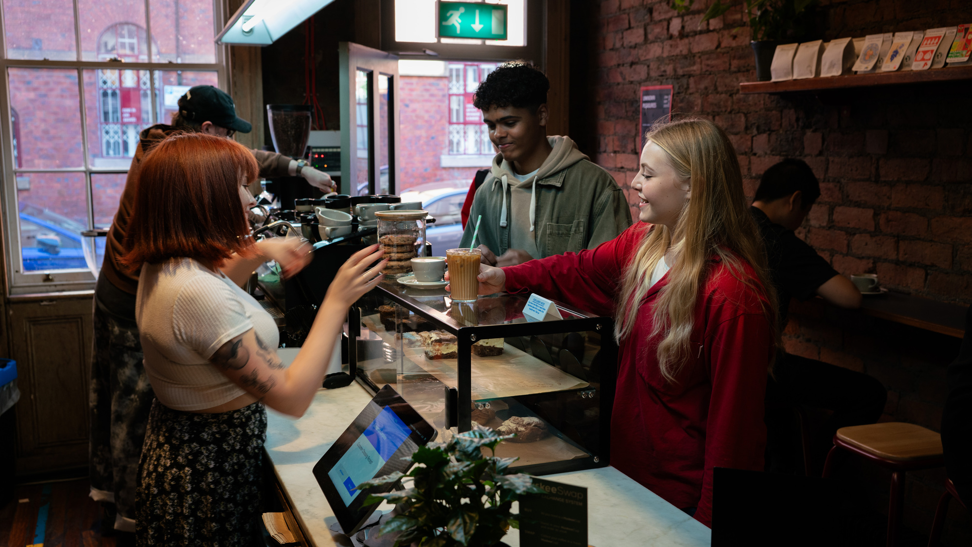 Barista serving a takeaway coffee to a customer inside a cozy café with exposed brick walls and wooden shelves displaying coffee bags. The counter features a glass display case with pastries, a small potted plant, and a payment terminal. A large window in the background lets in natural light, revealing a view of a red brick building outside.