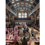 Wide view of a busy indoor craft market inside a grand historic hall with a high vaulted glass ceiling, ornate arches and large windows. Rows of vendor stalls fill the wooden parquet floor, displaying handmade artwork, prints, jewellery and accessories. Visitors browse displays throughout the hall. Sunlight streams through the windows, illuminating the market and highlighting the impressive architecture of the venue.