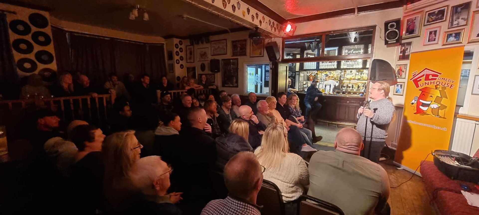 A lively comedy night in a cosy pub, with a seated audience watching a performer speaking into a microphone. The room is warmly lit, with framed pictures and vinyl records on the walls. A bright yellow banner for a comedy club stands beside the performer, and the bar area is visible in the background.