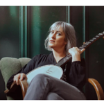 A person with short light-coloured hair sits in an upholstered armchair holding a banjo across their lap. Wearing a dark top and light trousers, the person looks towards the camera while resting one hand on the instrument’s neck. The background features dark green paneled walls, creating a moody, studio-style portrait with soft lighting and muted tones.