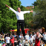 A performer balances on a very tall unicycle with arms outstretched during an outdoor event. Another performer in a red athletic outfit stands nearby, appearing to assist. Behind them, a large crowd watches, with colourful bunting, trees, and fairground stalls visible in the background on a sunny day.