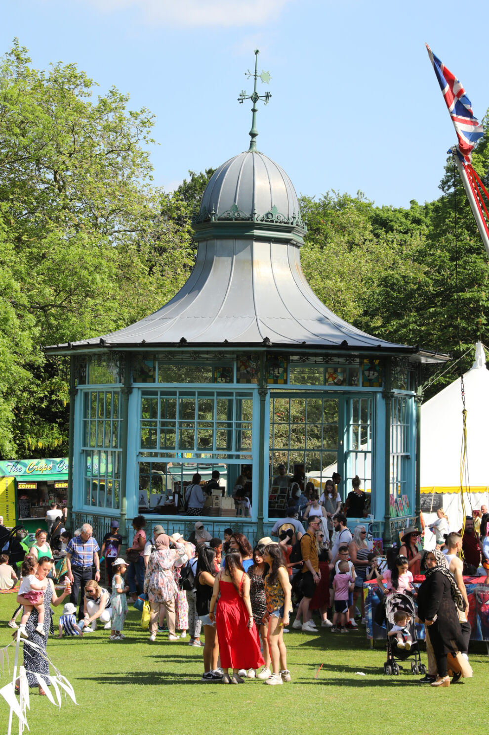 A decorative Victorian-style bandstand painted in blue stands in a park during a sunny outdoor event. People gather around and inside the structure, with groups chatting and walking on the grass. The background features lush green trees, a white marquee tent, and a tall flagpole with a Union Jack flag. The scene is lively, with festival activities taking place in a bright, open setting.