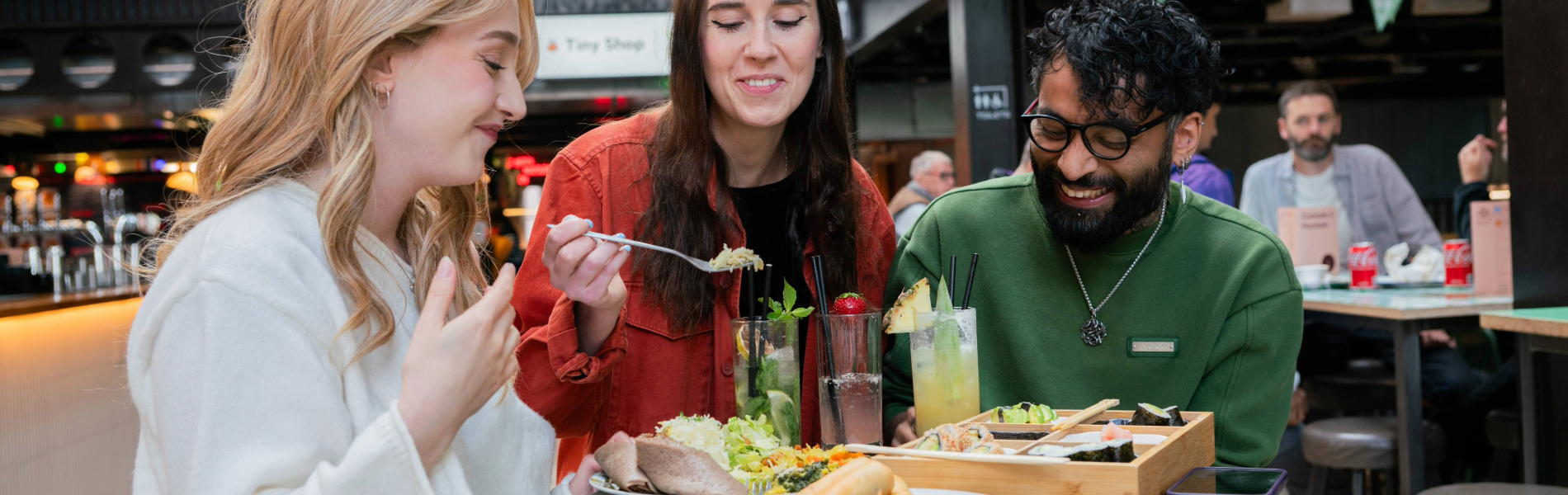 Three people seated at a round green table in an indoor food hall, sharing a meal. The table holds plates of tacos, salad, bread, and a wooden tray with assorted dishes. There are also tall glasses of colourful drinks garnished with fruit and herbs. In the background, green triangular banners hang overhead, and signs for “Oktoberfest” and “Baity” are visible, creating a lively, casual dining atmosphere.