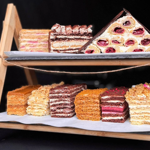 Selection of layered cakes and pastries displayed on a two-tier wooden stand against a dark background. The desserts include cream-filled sponge cakes, chocolate layered cakes and rolled pastries with a red fruit filling. The cakes are arranged on trays lined with white paper, showcasing a variety of colours, textures and decorative toppings.