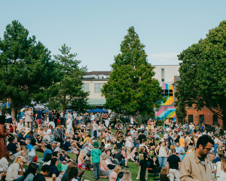 Festivalgoers sitting and socialising on grassy parkland at Devonshire Green during a busy outdoor music event.