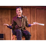 A person is sat on a high stool in a wood panelled room, smiling and gesticulating.
