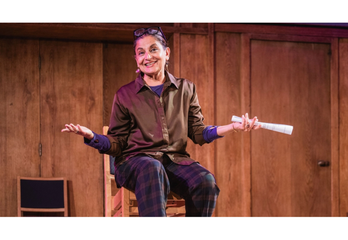 A person is sat on a high stool in a wood panelled room, smiling and gesticulating.