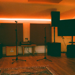 Music studio with warm ambient lighting, featuring two large speakers on stands, two microphone stands, and a flat-screen TV on the right. Acoustic panels are mounted on the back wall above a small wooden table holding audio equipment. An orange guitar amplifier sits on the floor to the left. The room has wooden flooring and a patterned rug in the foreground, creating a cosy, professional recording space.