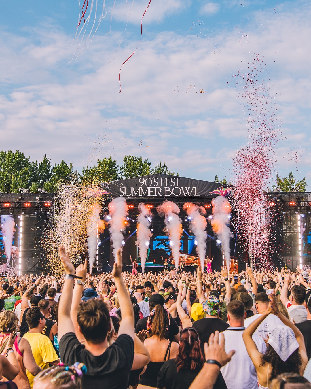 A lively festival crowd faces the “90’s Fest Summer Bowl” stage as smoke cannons shoot white plumes and colorful confetti fills the air. People have their hands raised, and the scene is bright and energetic with trees and blue sky in the background.