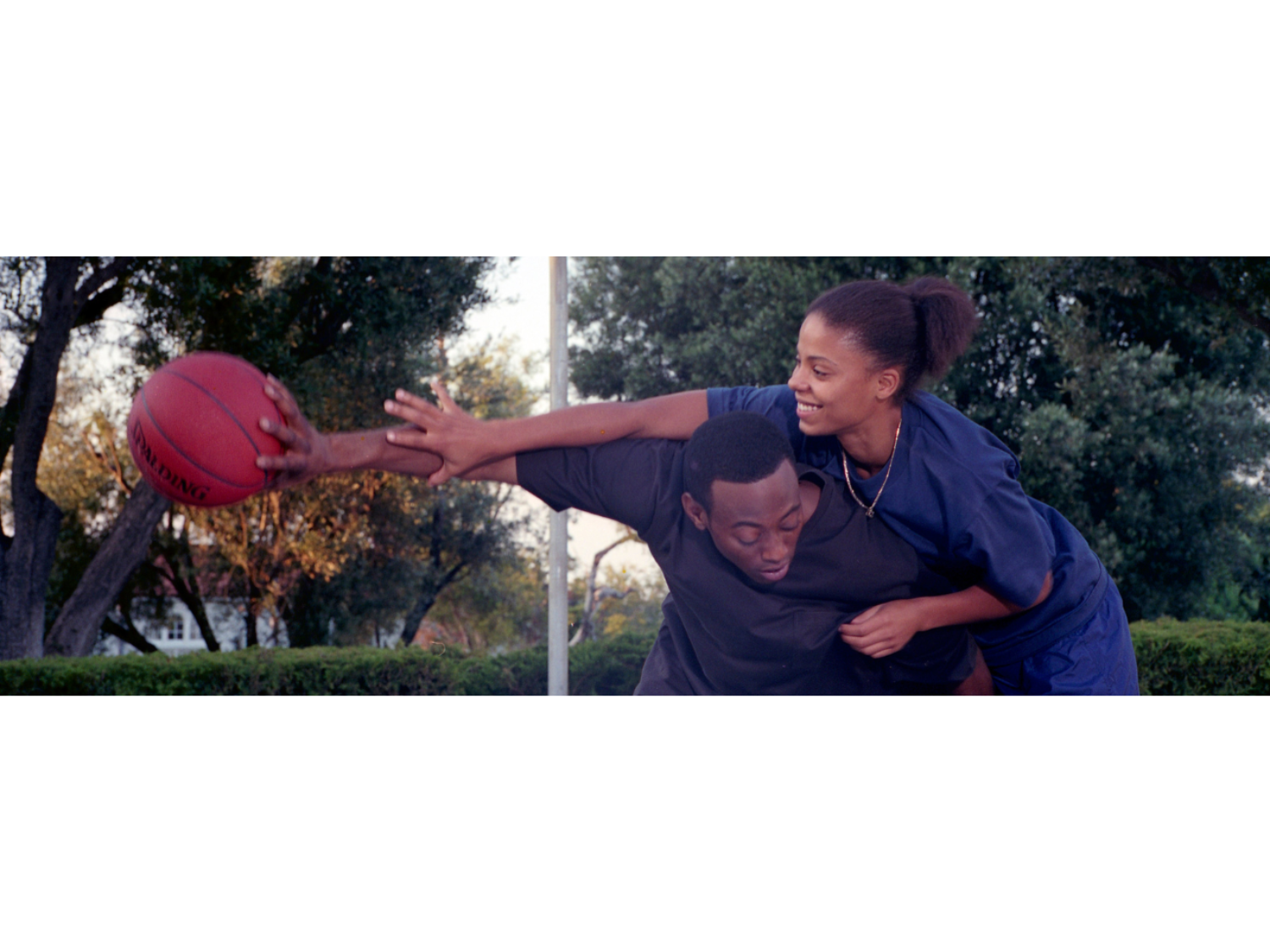 Two people playing basketball outdoors, surrounded by green trees and bushes. One person in a dark outfit holds a red basketball while leaning forward, as the other person in a blue sports outfit reaches over to block or steal the ball. The scene captures dynamic movement and physical interaction, with natural daylight and a park-like setting in the background.