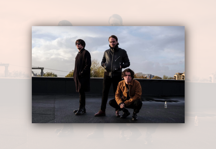 Three people pose on a rooftop or outdoor terrace under a cloudy sky. One stands wearing sunglasses and a long dark coat, another stands in a black leather jacket with hands in pockets, and a third crouches in a brown jacket and dark trousers. Buildings and trees are visible in the distance beyond a low wall. The image has a moody, urban promotional style, with soft evening light reflecting off the wet surface beneath them.