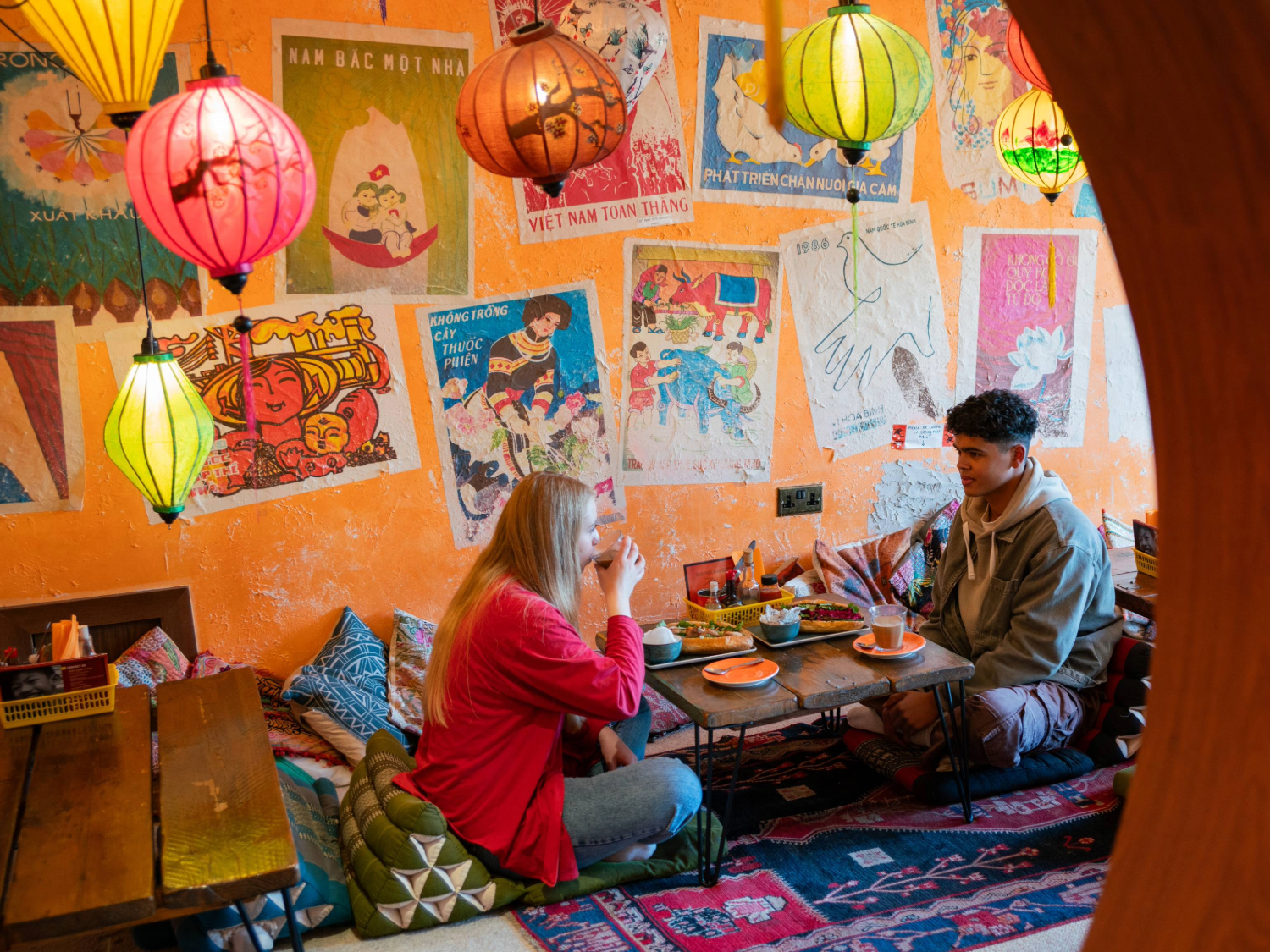 Two people are seated on cushions on the floor, at a low table, enjoying the meal. There are brightly coloured lanterns hanging from the ceiling. The orange walls are decorated with old and faded posters.