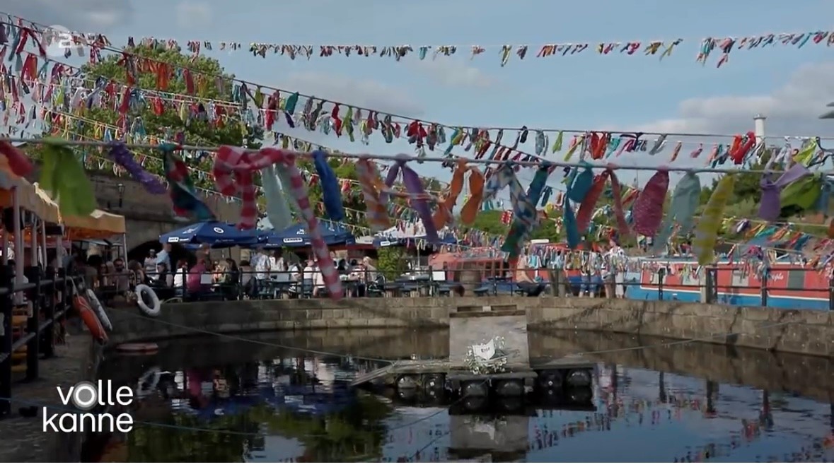 Outdoor canal-side seating area decorated with colorful fabric bunting strung across the space. People are sitting at tables under umbrellas, and narrowboats are moored along the water. The scene is bright and festive under a partly cloudy sky. Text on the bottom left reads ‘volle kanne.’