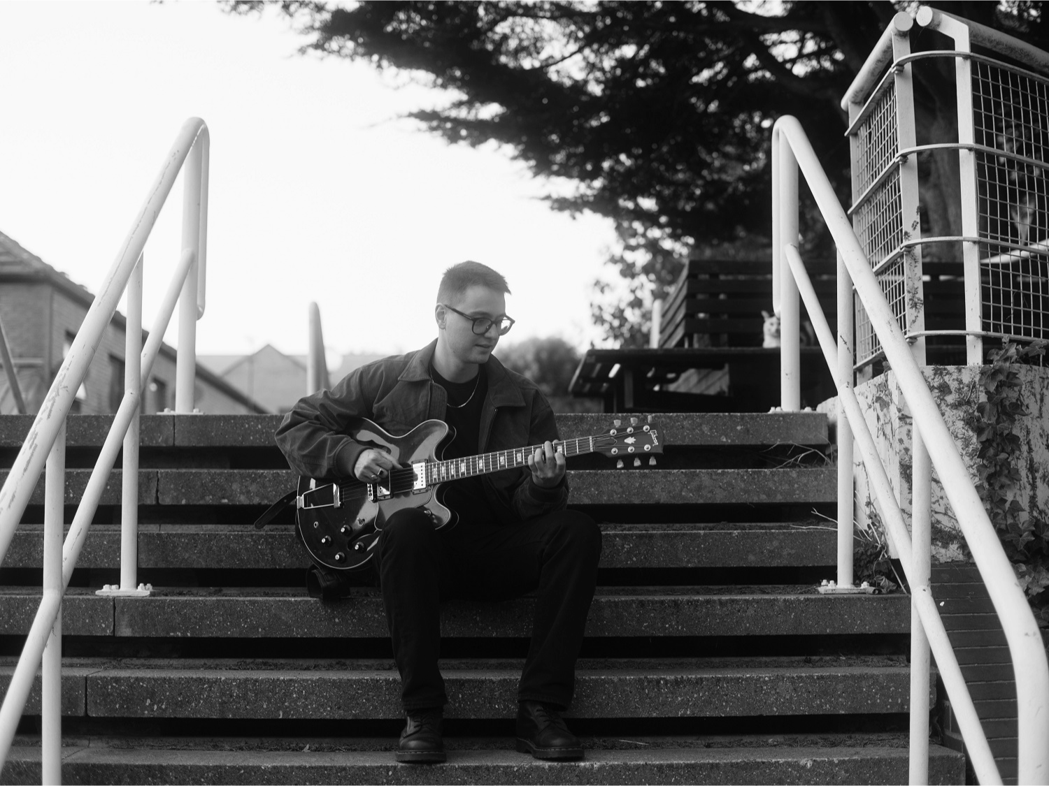 A person is seated on outdoor concrete steps, playing an electric guitar. The guitar has a semi-hollow body with a dark finish and visible f-holes. The individual is wearing a dark jacket and trousers, with both hands positioned on the guitar neck and body. White railings frame the steps on either side, and a tree with dense foliage is visible in the background along with a bench and part of a stone wall. The image is in black and white, creating a classic, moody atmosphere.