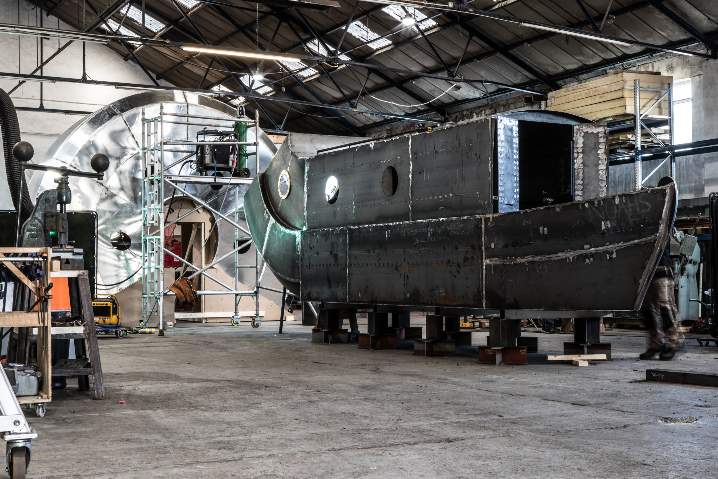 Interior of a large industrial workshop with high ceilings and metal beams, showing a partially constructed steel structure resembling a boat hull on supports. In the background, scaffolding surrounds a large circular metallic component, with tools and equipment scattered around the concrete floor.
