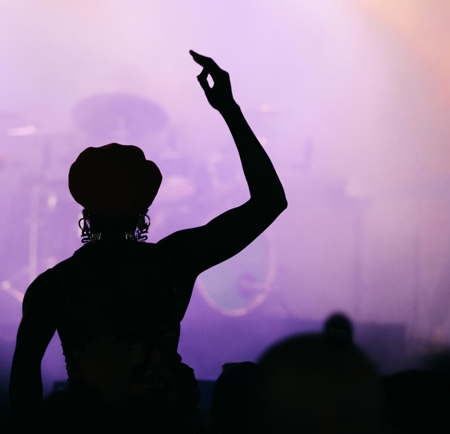 Silhouette of a person wearing a hat, raising one hand in front of a stage with purple and pink lighting and blurred musical instruments in the background.