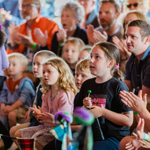 Children and adults sit closely together in an audience at a family-friendly indoor event. Several children in the foreground watch a performance, some clapping, while one child holds a green lollipop. A person stands at the left edge facing away from the camera. The background shows a packed crowd, many applauding. Bright, warm lighting and shallow focus emphasise the children, with the audience softly blurred behind them.