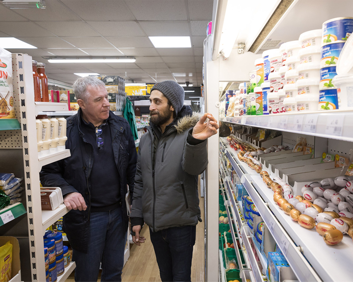 The inside of a food shop, with the owner and a customer standing between rows of shelves. 