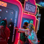 Three people playing an interactive arcade game called “Buzzer Beater” in a neon-lit venue. The game features a large vertical panel with multiple round buzzers and a digital score display reading “0550.” Bright signage on the machine includes phrases like “Green buzzers are great.” One person is actively playing, while another points at the panel, holding a drink. The setting is vibrant with colourful lights and a lively atmosphere.