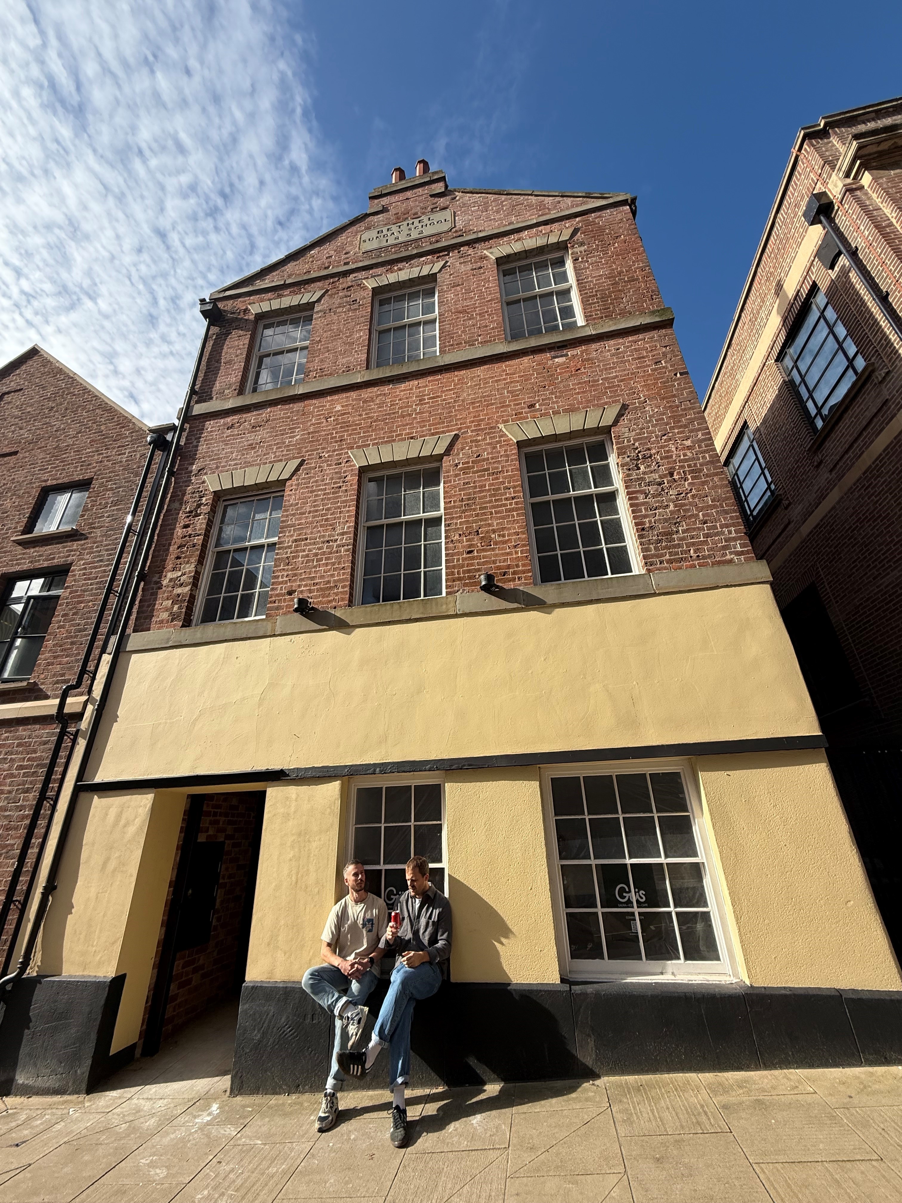 Tall brick building with three stories and a cream-colored ground floor facade. The upper floors have large rectangular windows with stone lintels, and the roofline features a small decorative gable with engraved text. Two people are sitting on the low ledge in front of the building, near a window with the word ‘Güs’ visible on the glass. The scene is lit by bright sunlight under a clear blue sky with wispy clouds.