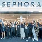 A large group of staff stand celebrating outside a brightly lit Sephora store during its opening event, holding ribbon and raising their arms in front of the storefront.