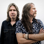 Two long-haired musicians pose against a plain light background. One stands facing the camera wearing a dark denim jacket, while the other stands slightly in front with arms folded, looking to the side and wearing a patterned short-sleeved shirt. The close-up portrait focuses on their contrasting poses and hairstyles, creating a simple, studio-style promotional image.