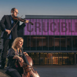 In Tudor Square, outside the Crucible Theatre, a woman plays a cello and a man plays a violin at dusk. 