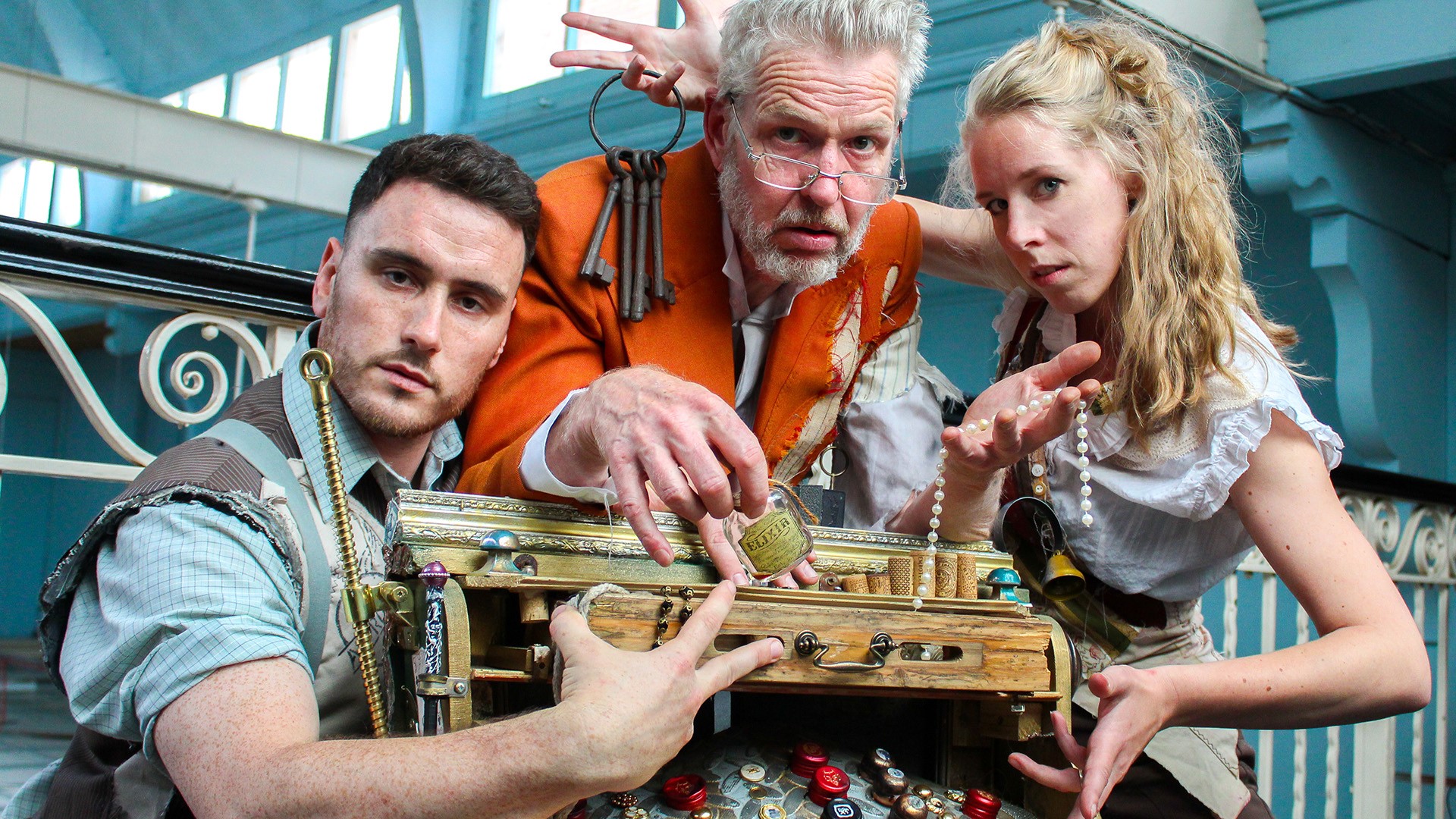 Three people dressed in steampunk-style costumes gathered around a decorative wooden box with brass fittings and assorted objects attached, including keys, locks, and bottle caps. The setting appears to be indoors with ornate railings and large windows in the background.