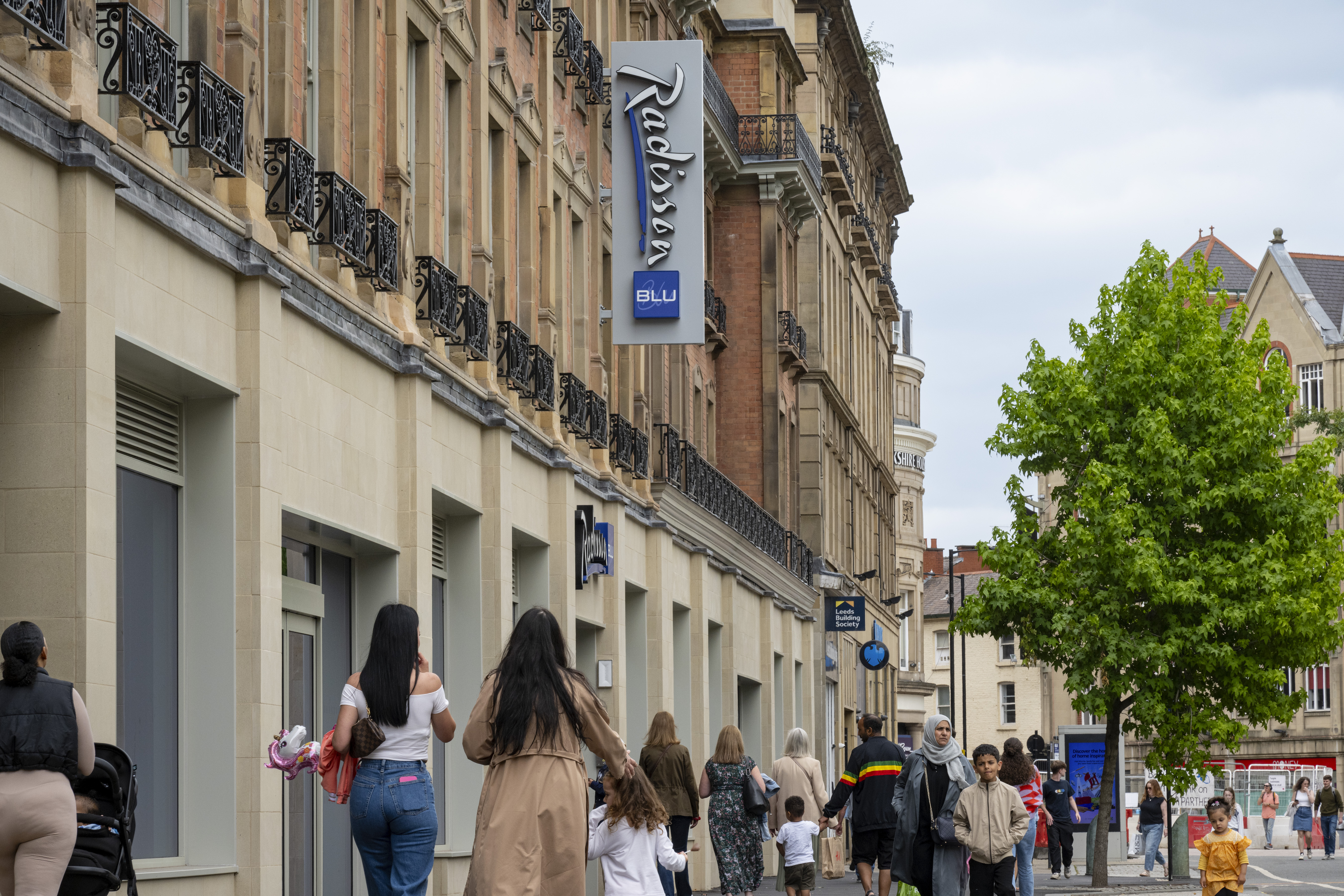 The exterior of the Radisson Blu Hotel on Pinstone Street in Sheffield city centre. The street is filled with people walking up and down. Above them hangs the Radisson Blu sign.